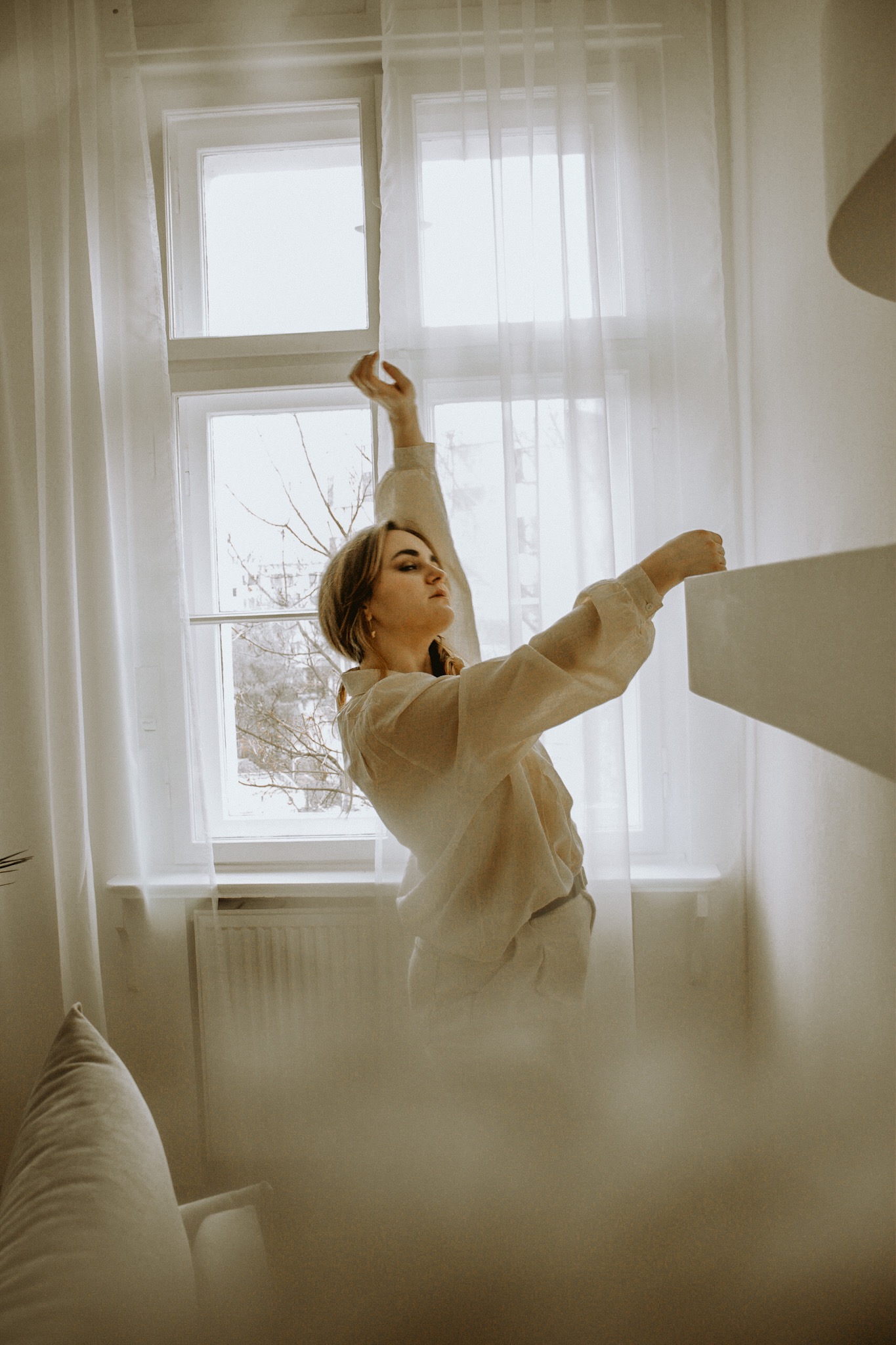 Kobieta w jasnej koszuli pozuje przy oknie z białymi zasłonami, delikatne światło podkreśla jej profil.