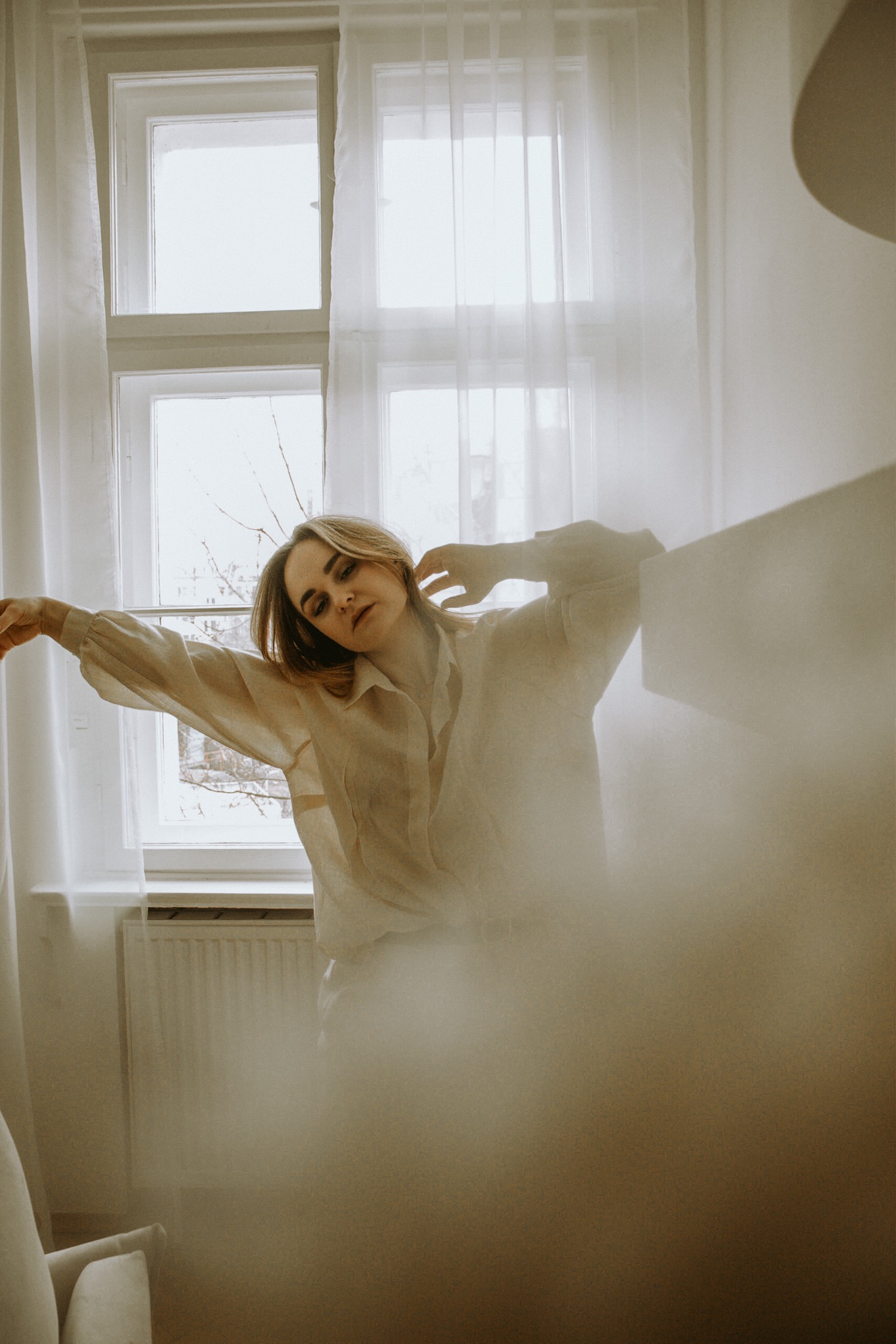 Kobieta w lnianej koszuli pozuje przy oknie z białymi zasłonami, delikatne światło pada na twarz, artystyczne rozmycie w dolnej części kadru.