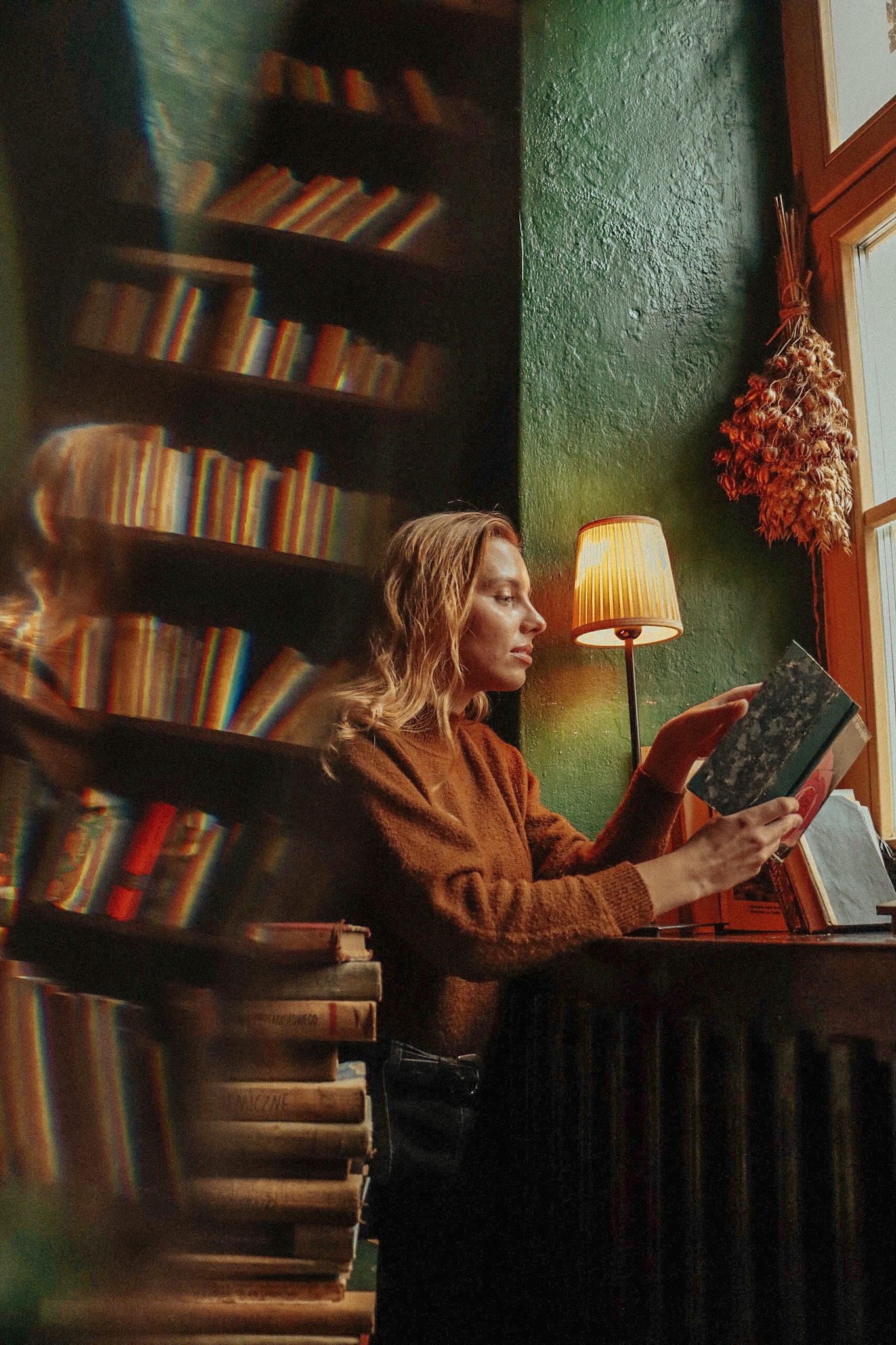 Kobieta w swetrze czyta książkę w klimatycznym wnętrzu biblioteki z zielonymi ścianami, oświetlonym ciepłym światłem lampki nocnej, obok okna z suszonymi kwiatami.