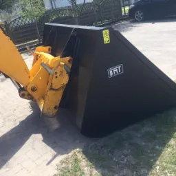 Czarny, stalowy osprzęt koparki z żółtym ramieniem montażowym, widoczne ślady użytkowania i logo DMT.