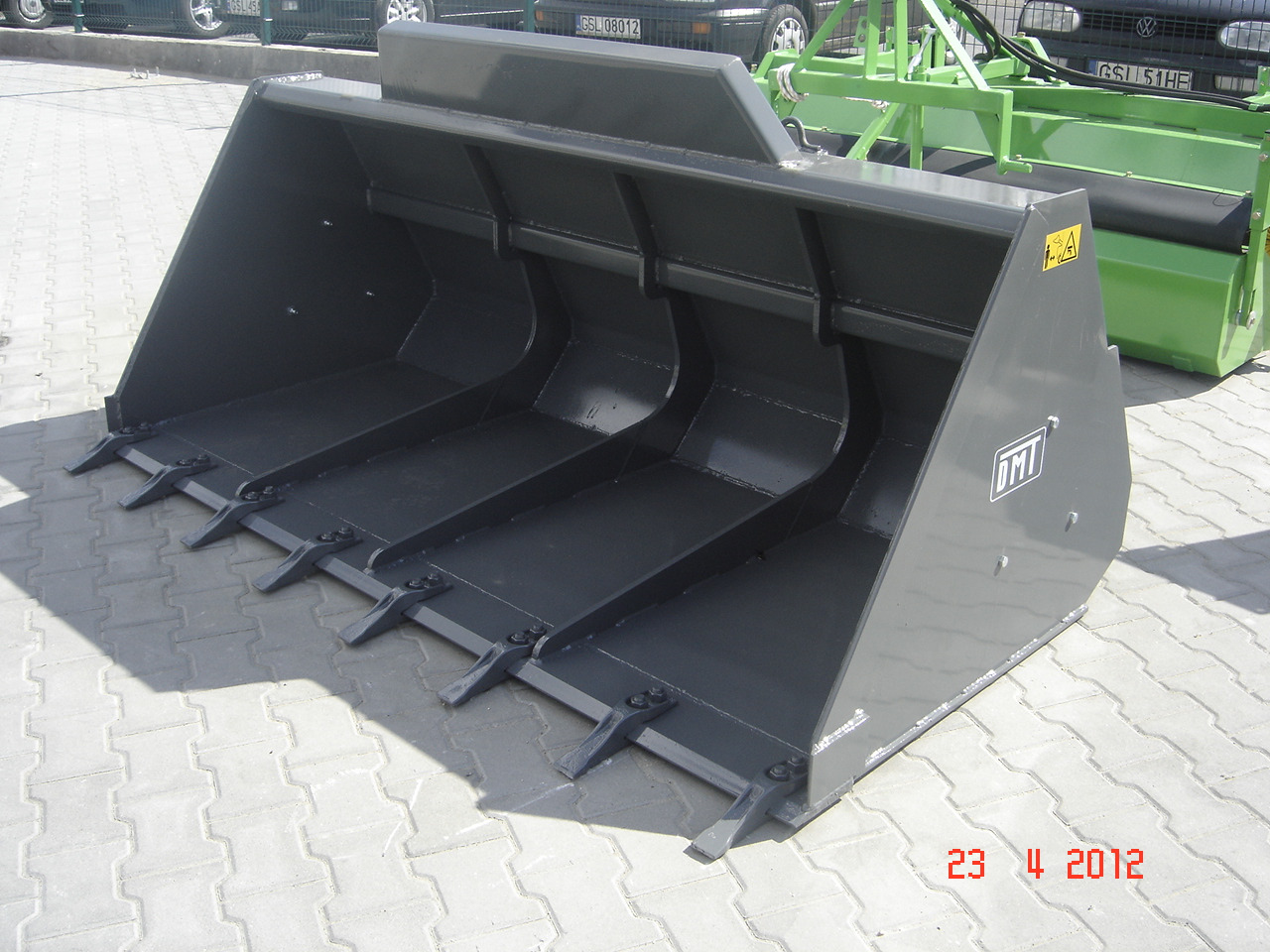 Szara, metalowa łyżka do ładowarki z zębami, widoczna na tle brukowanej powierzchni i fragmentu zielonej maszyny rolniczej, z naklejką ostrzegawczą i logo DMT.