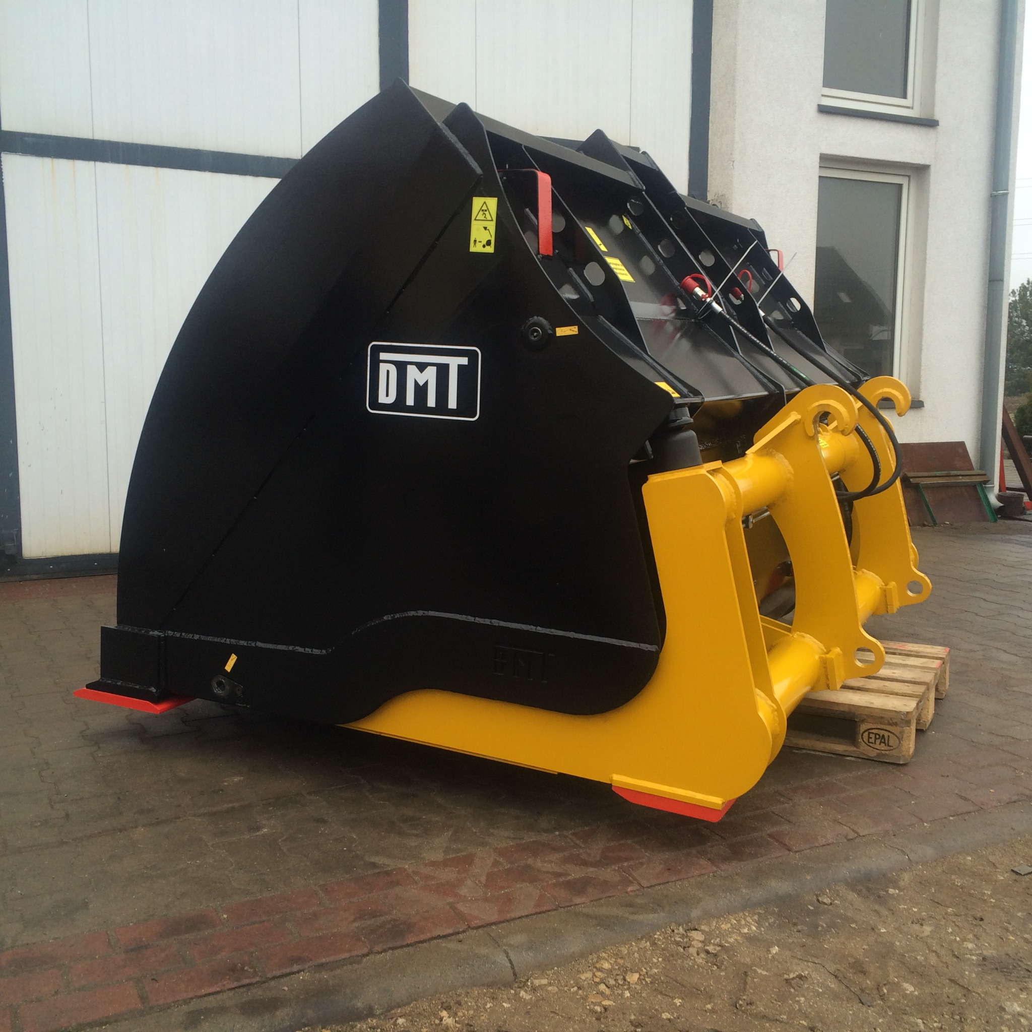 Nowa, czarno-żółta łyżka do ładowarki, z logo DMT, umieszczona na palecie EPAL przed budynkiem.