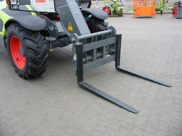 Szare widły do podnoszenia przymocowane do maszyny rolniczej, widoczne opony z czerwonymi felgami i ostrzeżenie na ramieniu.