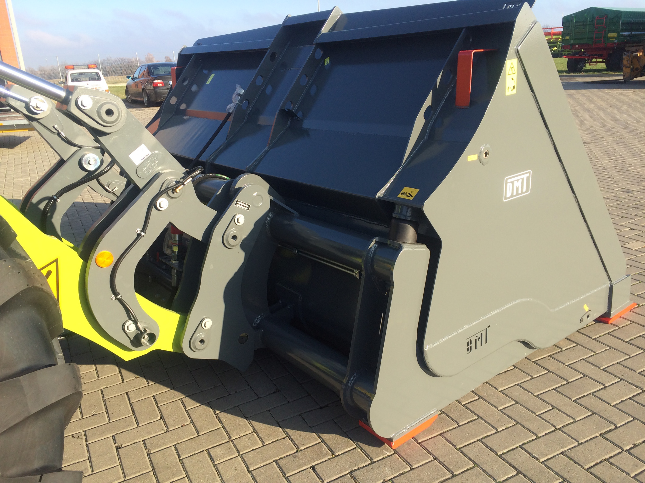 Szaro-grafitowy osprzęt do ładowarki teleskopowej, widoczne mocowania i logo DMT, na kostce brukowej.