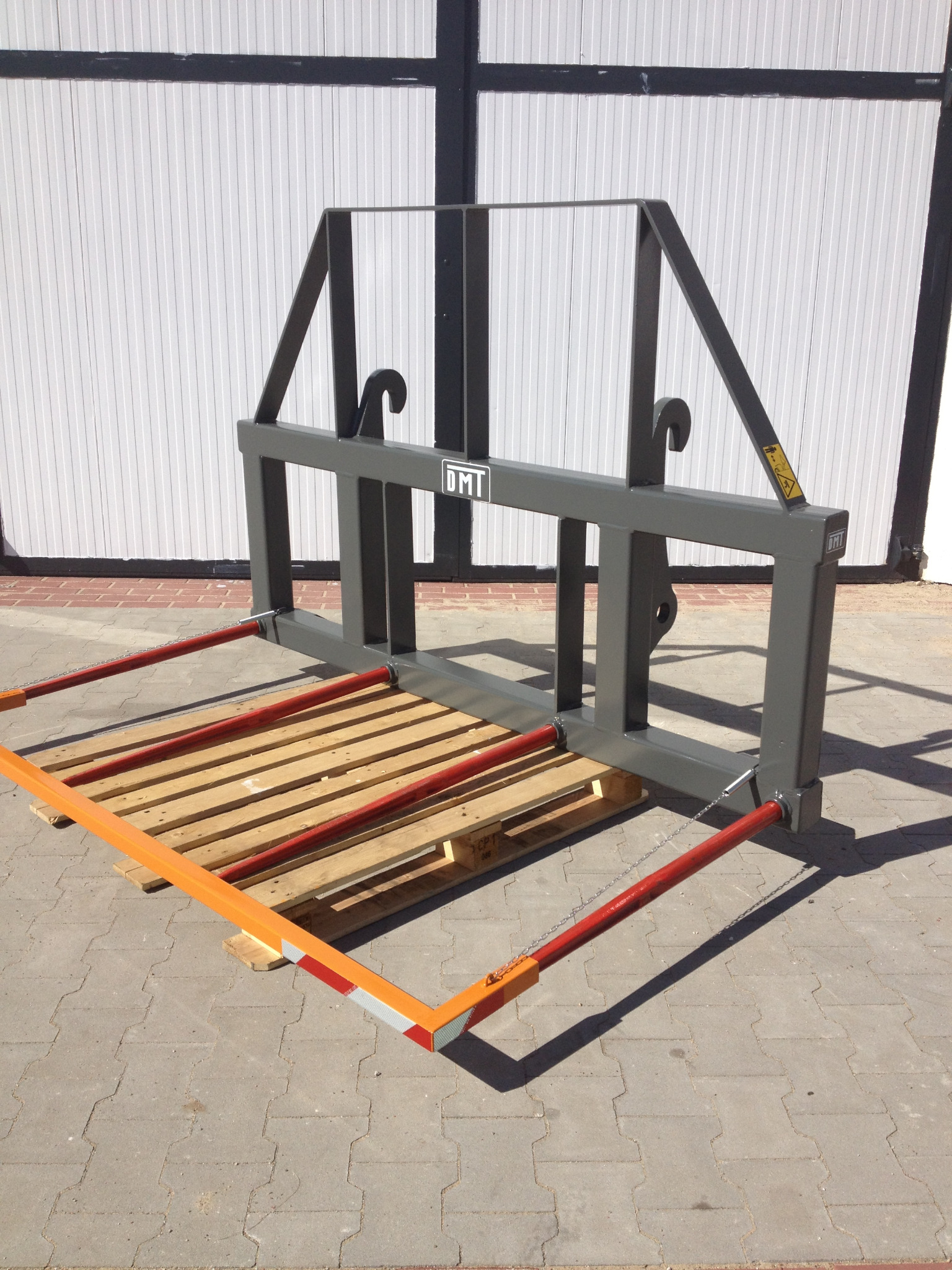 Szary, metalowy chwytak do palet z logo DMT, z pomarańczowo-czerwonymi elementami zabezpieczającymi, umieszczony na palecie na tle szarej kostki brukowej i białej hali.