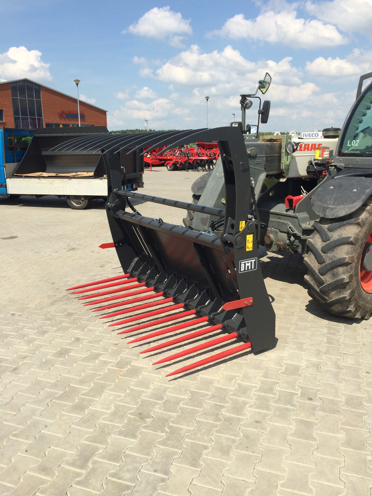 Czerwone widły do obornika zamontowane na ładowarce teleskopowej, widoczne na tle placu wyłożonego kostką brukową.