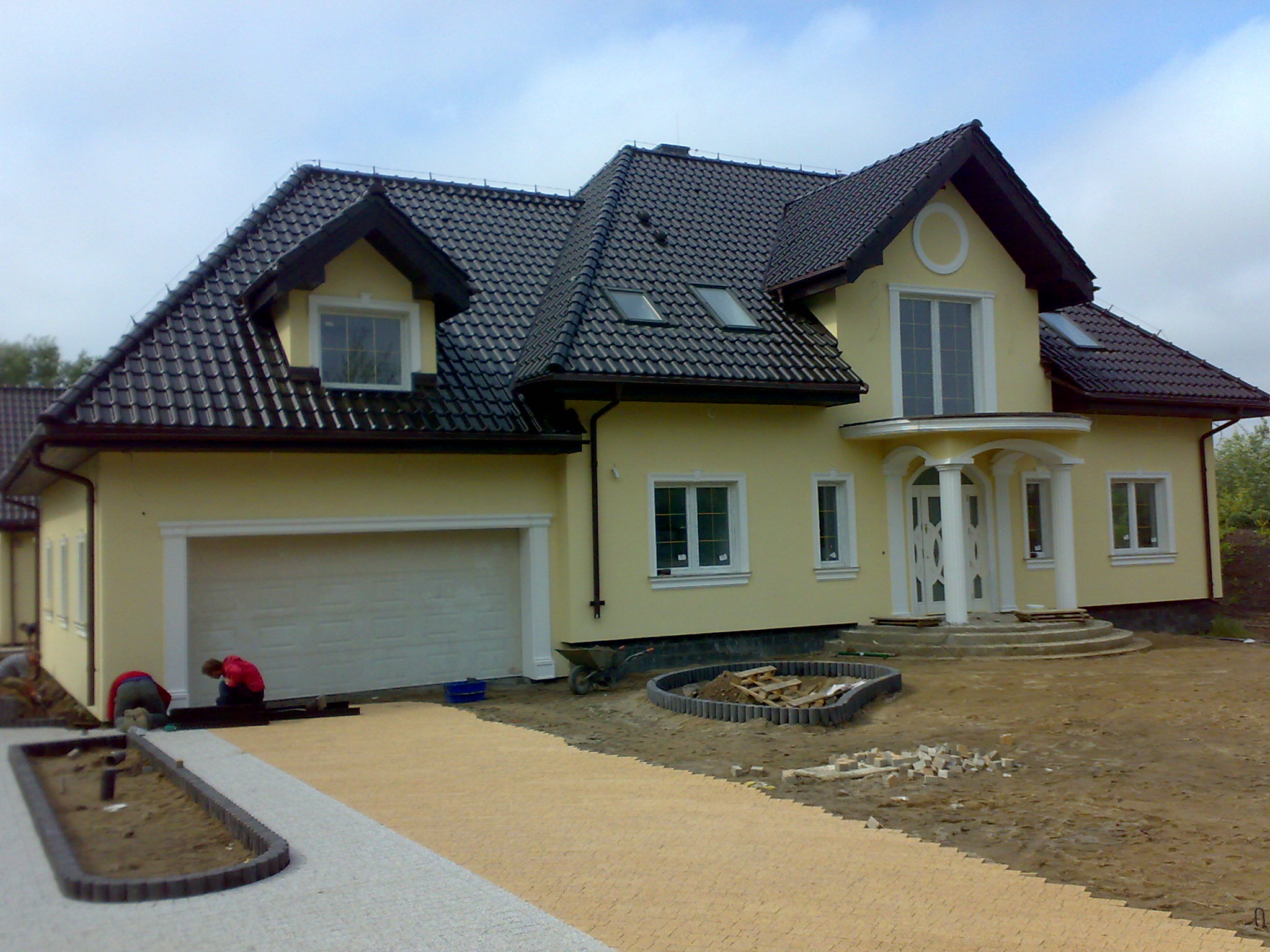 Żółta elewacja domu jednorodzinnego z ciemną dachówką, widoczni pracownicy układający kostkę brukową na podjeździe i obrzeża rabaty.
