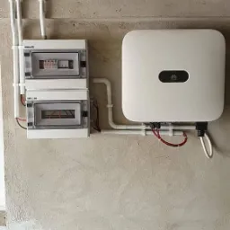 Biały inwerter z logo Huawei oraz dwie białe skrzynki bezpiecznikowe zamontowane na surowej, betonowej ścianie z widocznymi kablami i rurkami instalacyjnymi.