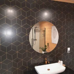 Nowoczesna łazienka z czarną ścianą pokrytą geometrycznym wzorem złotych sześcianów, okrągłe lustro odbijające prysznic i drewniane panele, biała umywalka nablatowa z miedzianą baterią.