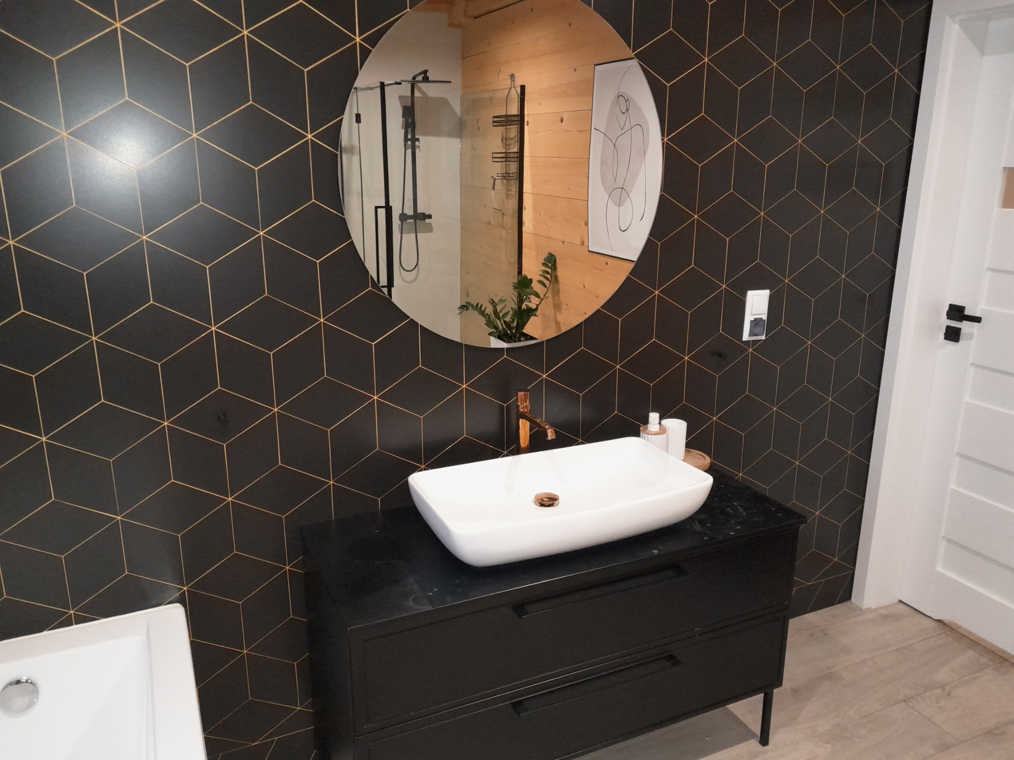 Nowoczesna łazienka z czarną ścianą w geometryczne wzory, okrągłe lustro odbijające kabinę prysznicową, biała umywalka nablatowa na czarnej szafce z szufladami i złotą armaturą.