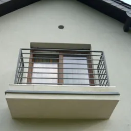 Zewnętrzny balkon z szarą, metalową balustradą z poziomych pręt&oacute;w, zamontowany na betonowej płycie, widoczny na tle okna i ściany budynku.