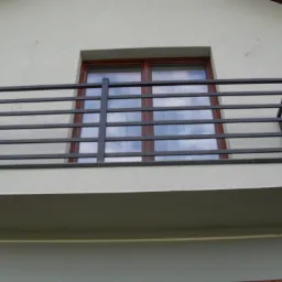 Czarna, pozioma balustrada balkonowa z metalowych profili na tle okna z odbijającym się niebem.