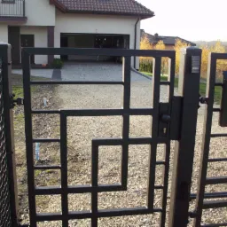 Otwarta metalowa brama z geometrycznym wzorem, skrzynka na listy z symbolem poczty, dom z otwartym garażem w tle.