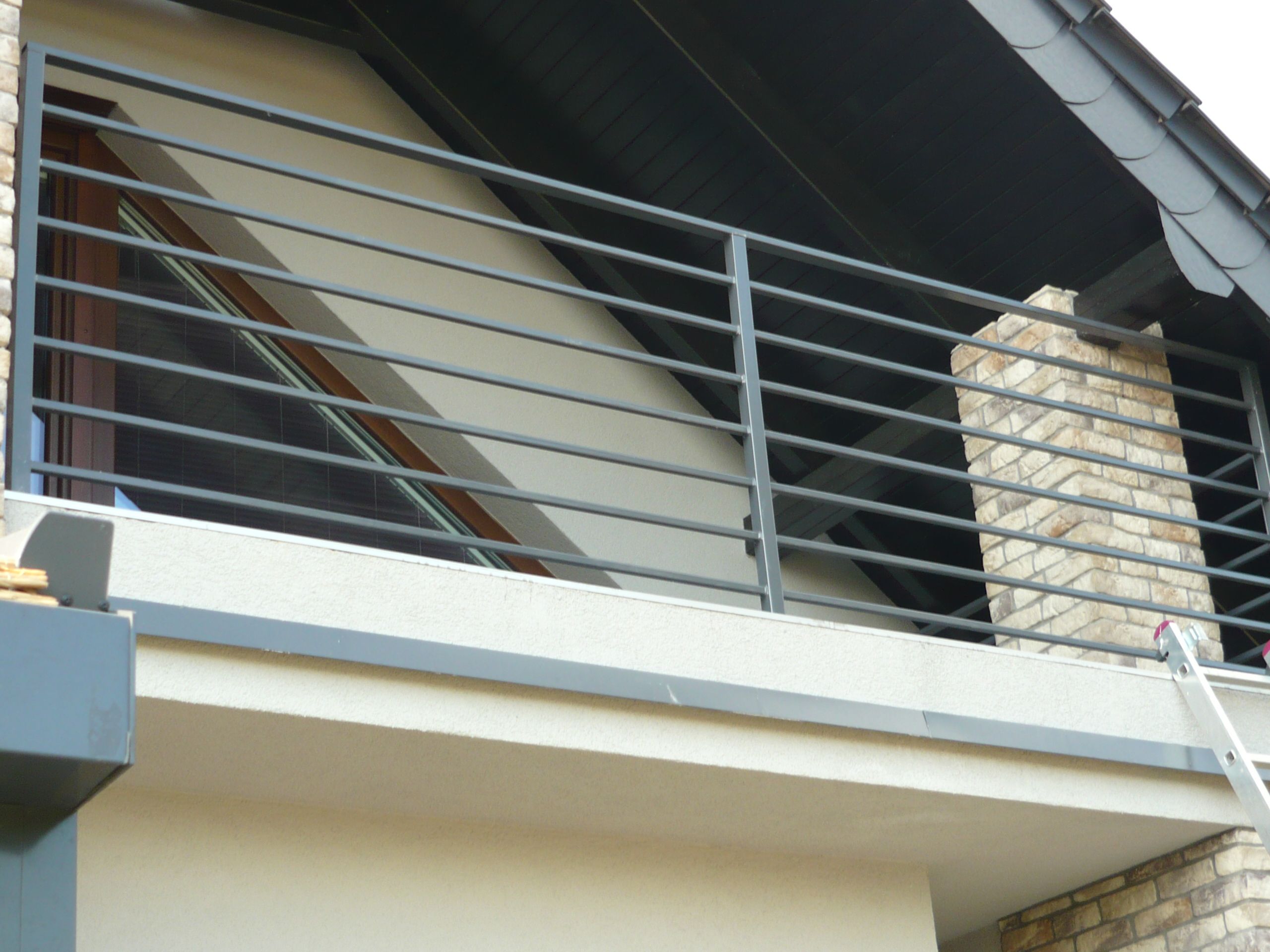 Szara, pozioma balustrada balkonowa z metalowych prętów na tle jasnej ściany i fragmentu dachu pokrytego ciemną dachówką, z fragmentem komina obłożonego cegłą po prawej stronie.