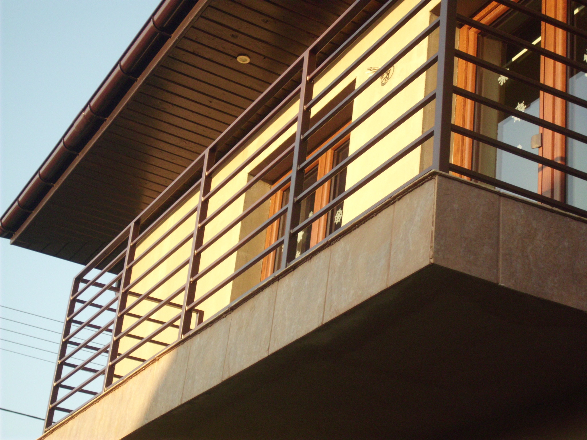 Kątowe ujęcie balkonu z metalową, poziomą balustradą w kolorze ciemnego brązu, na tle beżowej elewacji budynku z drewnianymi oknami. Widoczny fragment dachu z brązową podbitką.