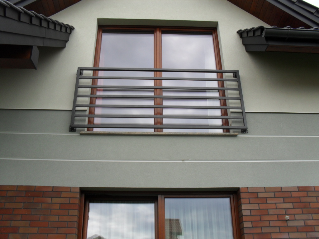 Szara, pozioma balustrada balkonowa z metalowych profili, zamontowana przed oknem na elewacji domu z ceglaną podmurówką.