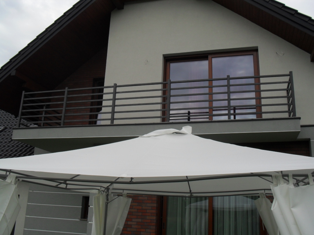 Szara elewacja budynku z balkonem zabezpieczonym poziomą, metalową balustradą w Sączowie, widoczna spod białego namiotu ogrodowego.