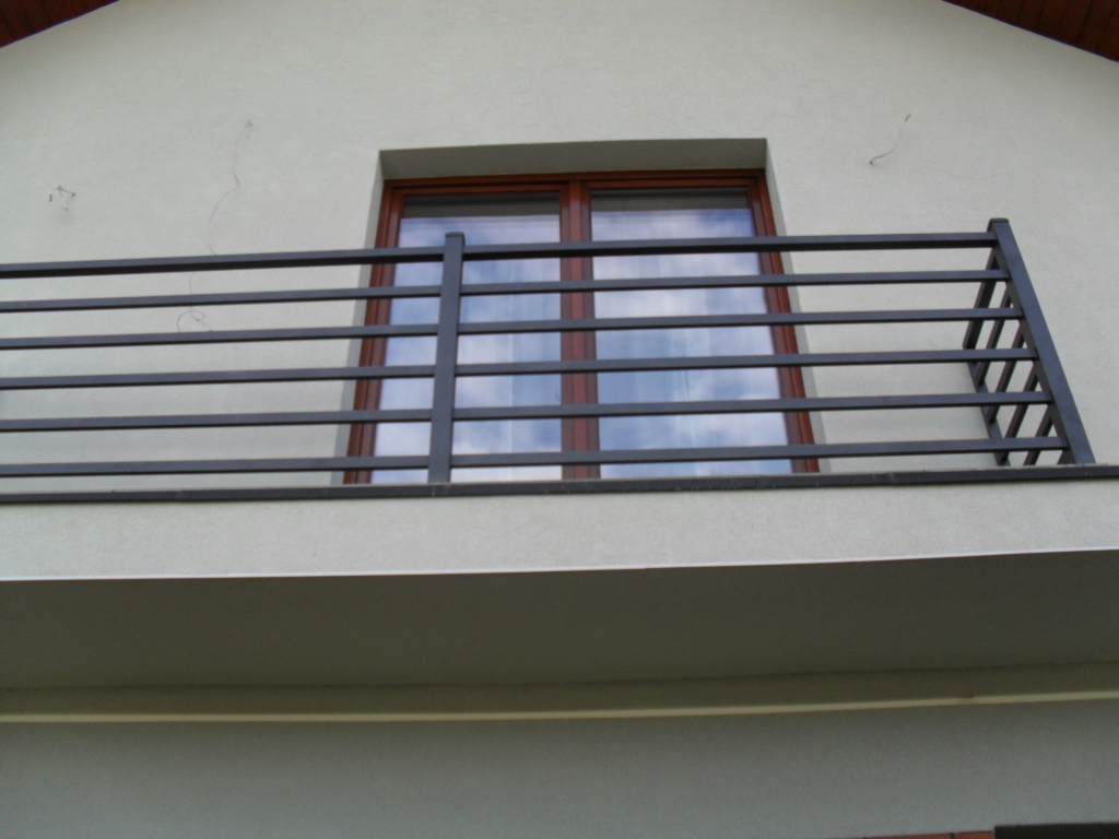 Czarna, pozioma balustrada balkonowa z metalowych profili na tle okna z odbijającym się niebem.