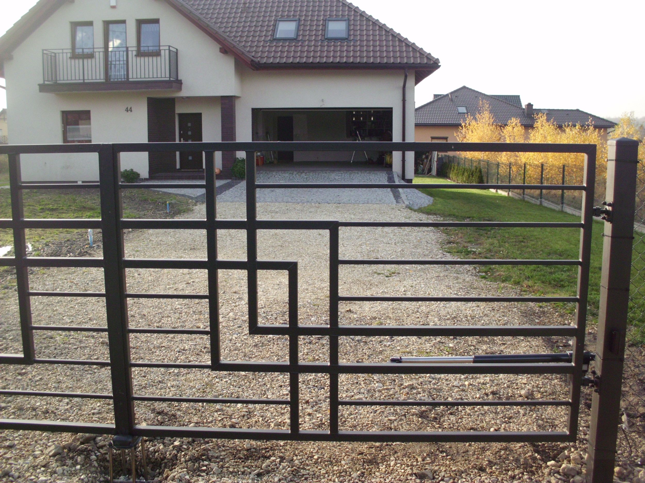 Nowoczesna, metalowa brama wjazdowa z geometrycznym wzorem, widoczny automat do otwierania, na podjeździe z jasnego żwiru, w tle dom jednorodzinny z garażem.