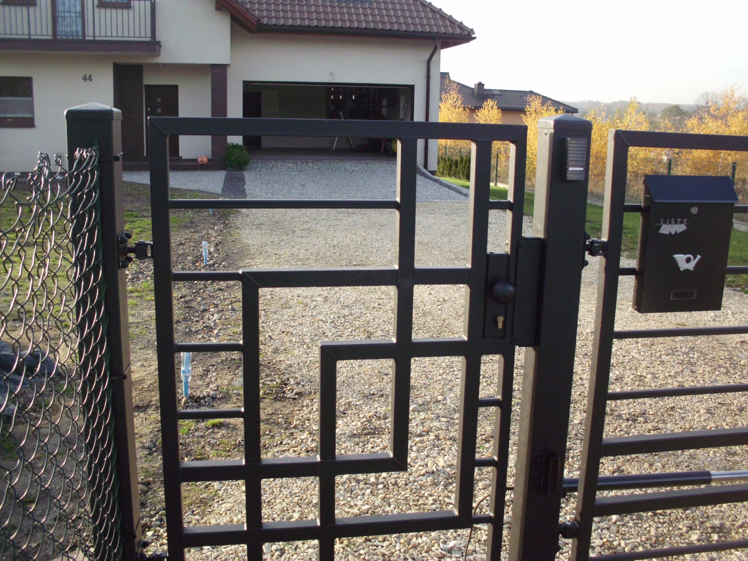 Otwarta metalowa brama z geometrycznym wzorem, skrzynka na listy z symbolem poczty, dom z otwartym garażem w tle.