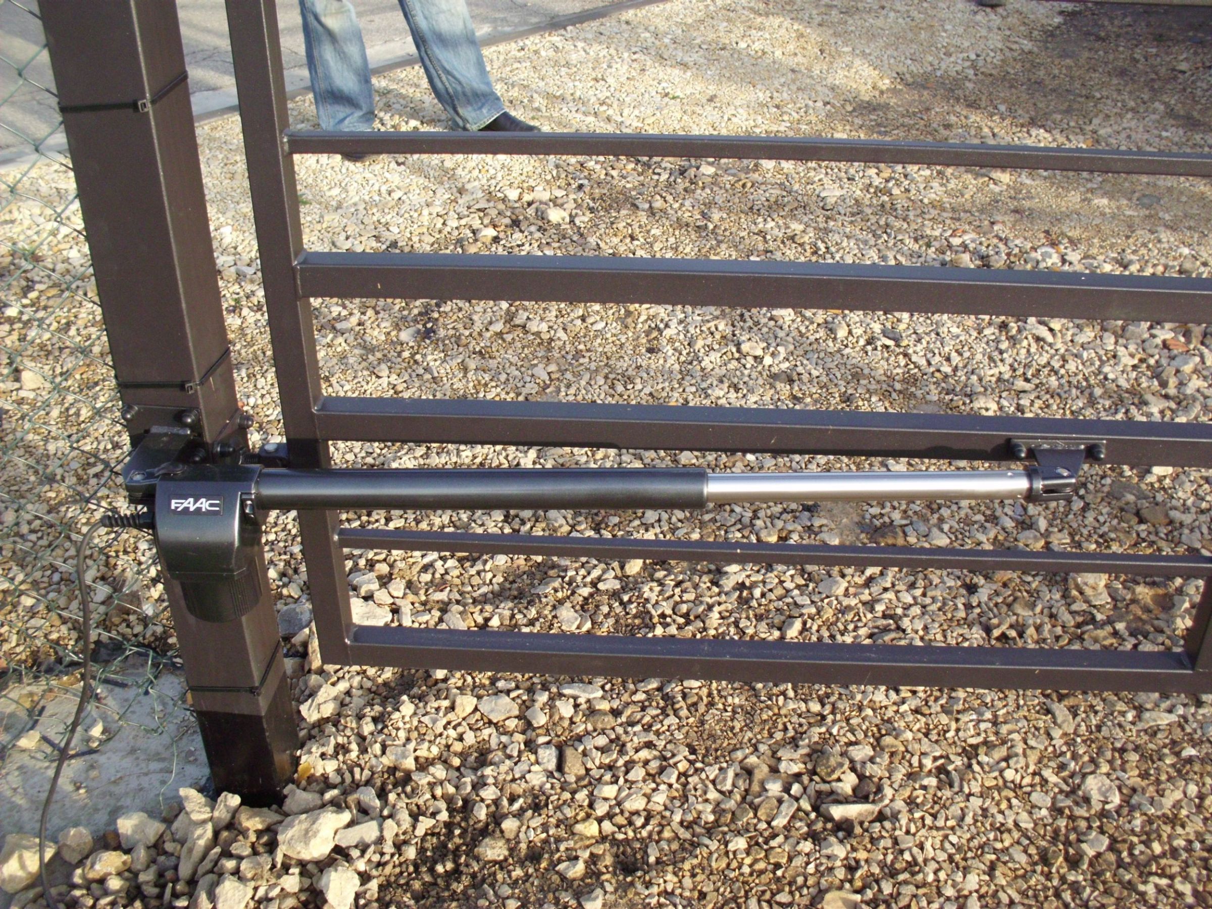 Fragment automatycznej bramy wjazdowej z siłownikiem FAAC, widoczny na tle żwirowego podłoża i fragmentu ogrodzenia z siatki.