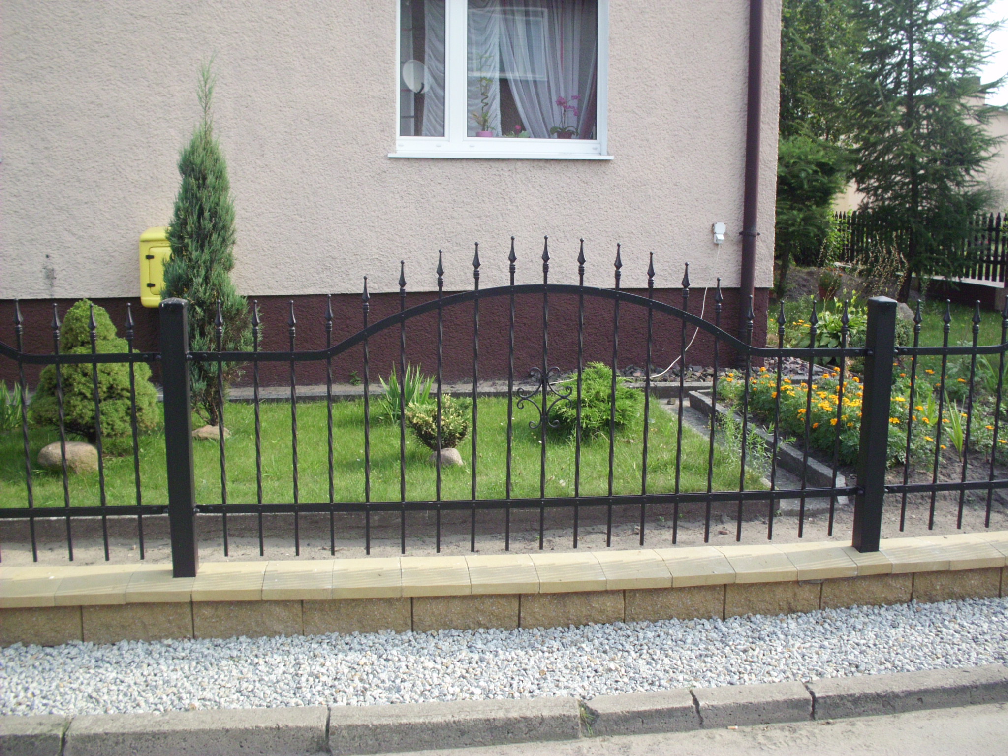 Czarne, metalowe ogrodzenie z ozdobnymi grotami na podeście z beżowej kostki, oddzielające trawnik z roślinami od chodnika wysypanego białym żwirem, na tle beżowego budynku z oknem.