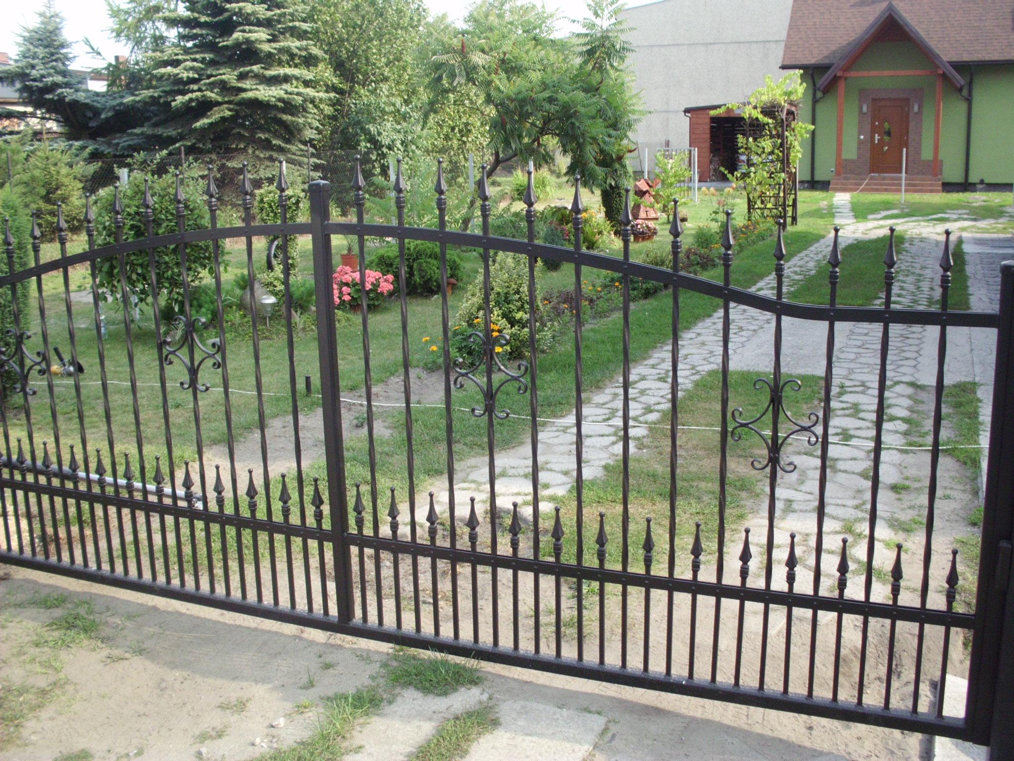 Otwarta, metalowa brama wjazdowa z dekoracyjnymi elementami, prowadząca do domu z zieloną elewacją i ścieżką z kamiennych płyt, widok z zewnątrz.