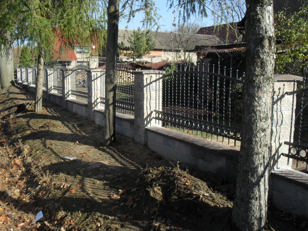 Długie ogrodzenie z metalowymi przęsłami i betonowymi słupkami, z widocznymi drzewami wzdłuż linii ogrodzenia, na tle zabudowań gospodarczych.