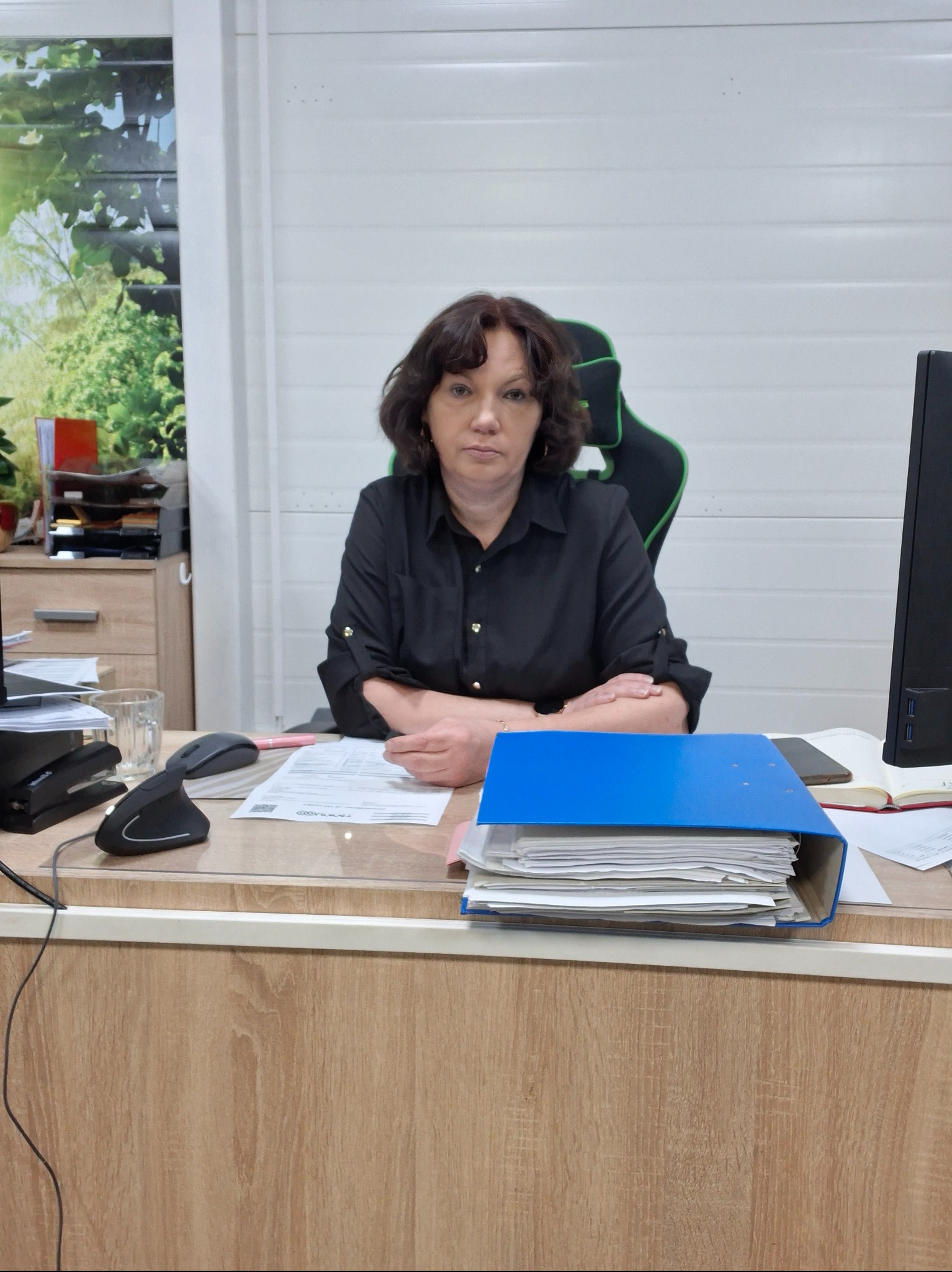 Kobieta siedzi za biurkiem z niebieskim segregatorze, dokumentami i telefonem. Widoczny monitor, myszka i elementy wyposażenia biura.