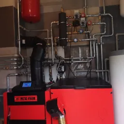 Czerwono-czarny piec Metal-Fach z podajnikiem ślimakowym i widoczną instalacją hydrauliczną, w pomieszczeniu kotłowni.