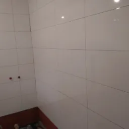 Łazienka w trakcie remontu: białe, błyszczące płytki na ścianach, częściowo wykończona podłoga z widocznym ogrzewaniem podłogowym, ściany częściowo zabezpieczone czerwoną farbą, widoczne przyłącza...