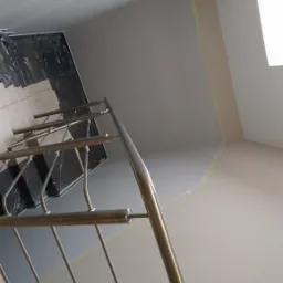 Świeżo pomalowane ściany klatki schodowej z metalową balustradą, zabezpieczone taśmą malarską przy suficie, z widocznymi schodami zakrytymi folią ochronną.