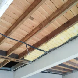 Fragment sufitu z drewnianymi belkami, częściowo ocieplony żółtą wełną mineralną, widoczne przewody elektryczne i fragment ściany w trakcie prac wykończeniowych.