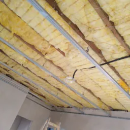 Ocieplenie poddasza z widoczną wełną mineralną między drewnianymi belkami i metalowymi profilami, przygotowane do montażu płyt gipsowo-kartonowych, drabina w rogu pomieszczenia.