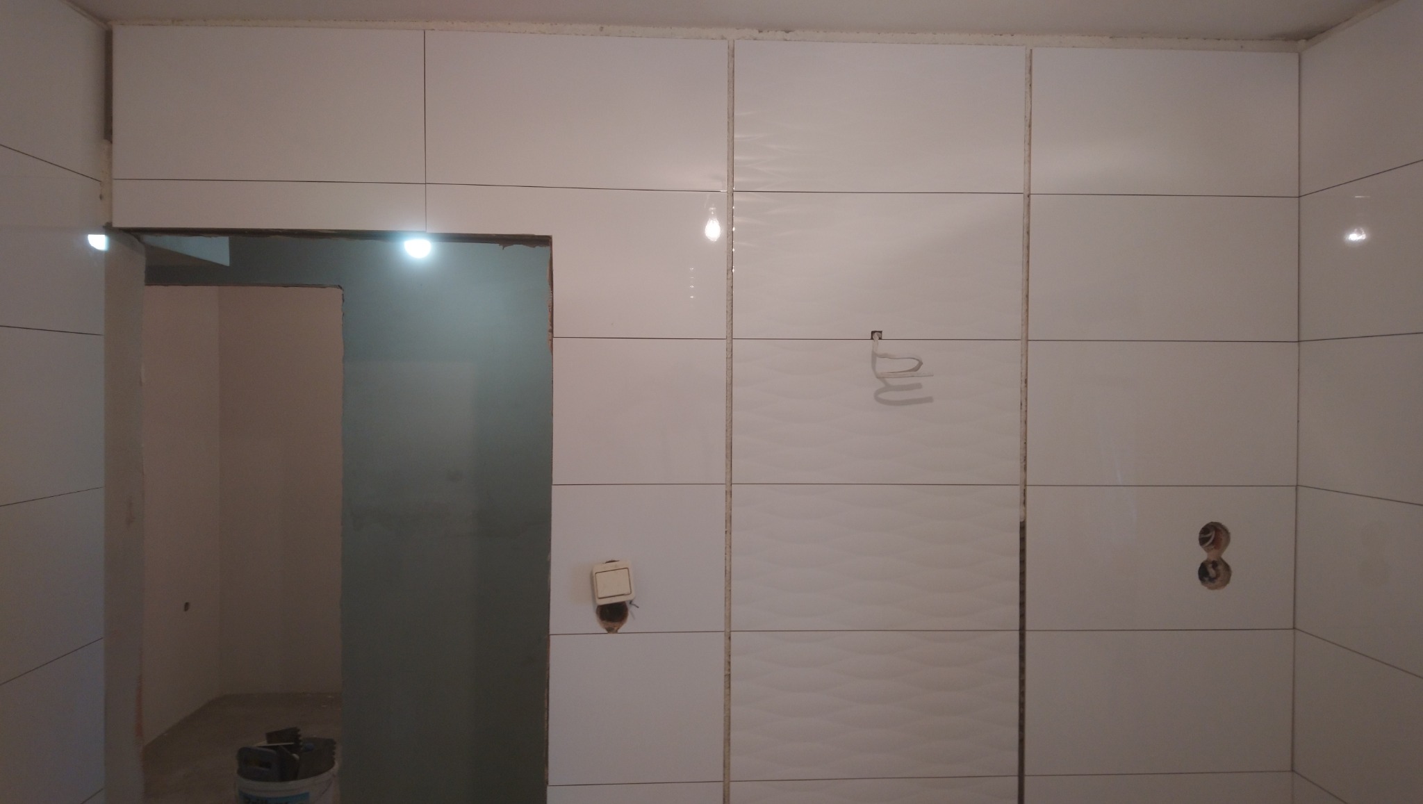 Ściana z białych, błyszczących płytek ceramicznych w trakcie układania, widoczne otwory na gniazdka elektryczne i włącznik światła, częściowo wykończone pomieszczenie z surowymi ścianami w tle.