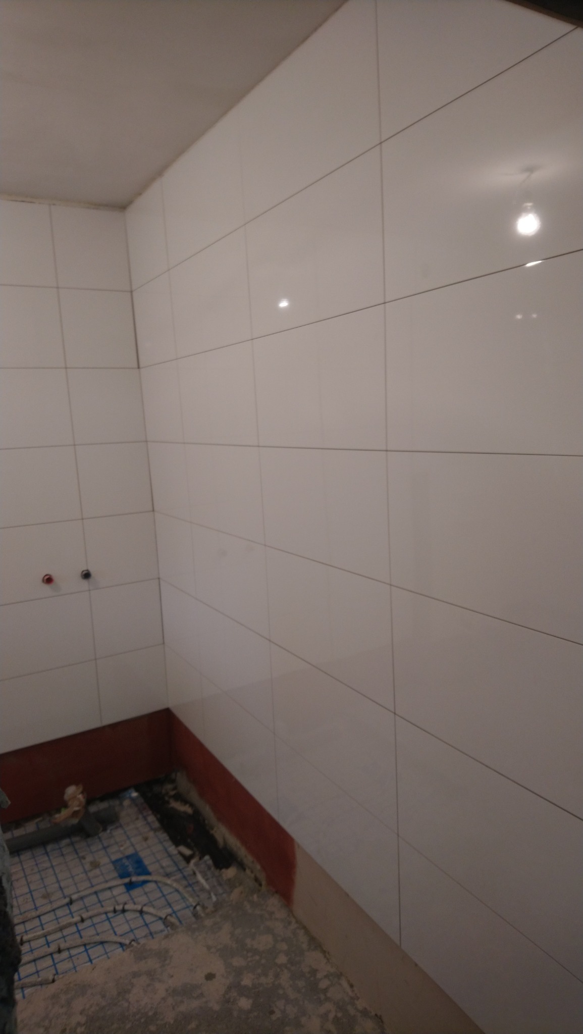 Łazienka w trakcie remontu: białe, błyszczące płytki na ścianach, częściowo wykończona podłoga z widocznym ogrzewaniem podłogowym, ściany częściowo zabezpieczone czerwoną farbą, widoczne przyłącza...