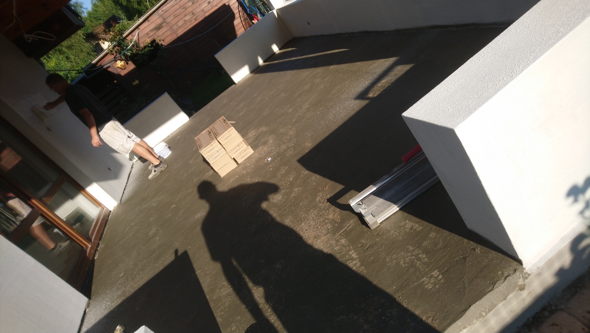 Prace wykończeniowe na tarasie: mężczyzna maluje ścianę na biało, obok leżą kartony i narzędzie do cięcia płytek, widoczny cień osoby robiącej zdjęcie.