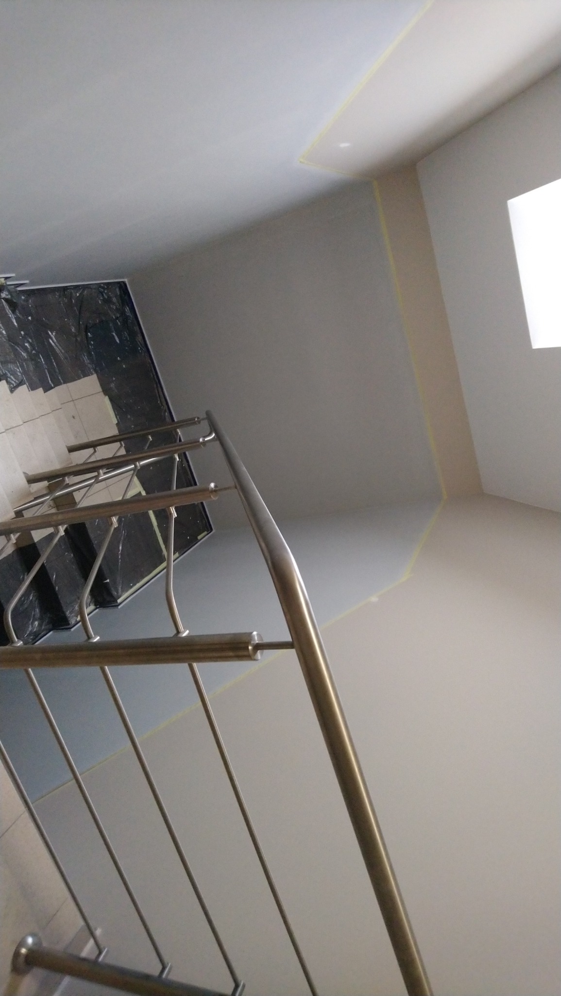 Świeżo pomalowane ściany klatki schodowej z metalową balustradą, zabezpieczone taśmą malarską przy suficie, z widocznymi schodami zakrytymi folią ochronną.