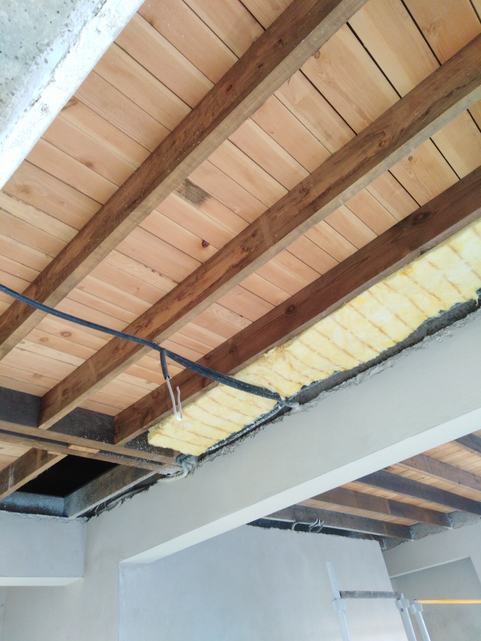 Fragment sufitu z drewnianymi belkami, częściowo ocieplony żółtą wełną mineralną, widoczne przewody elektryczne i fragment ściany w trakcie prac wykończeniowych.