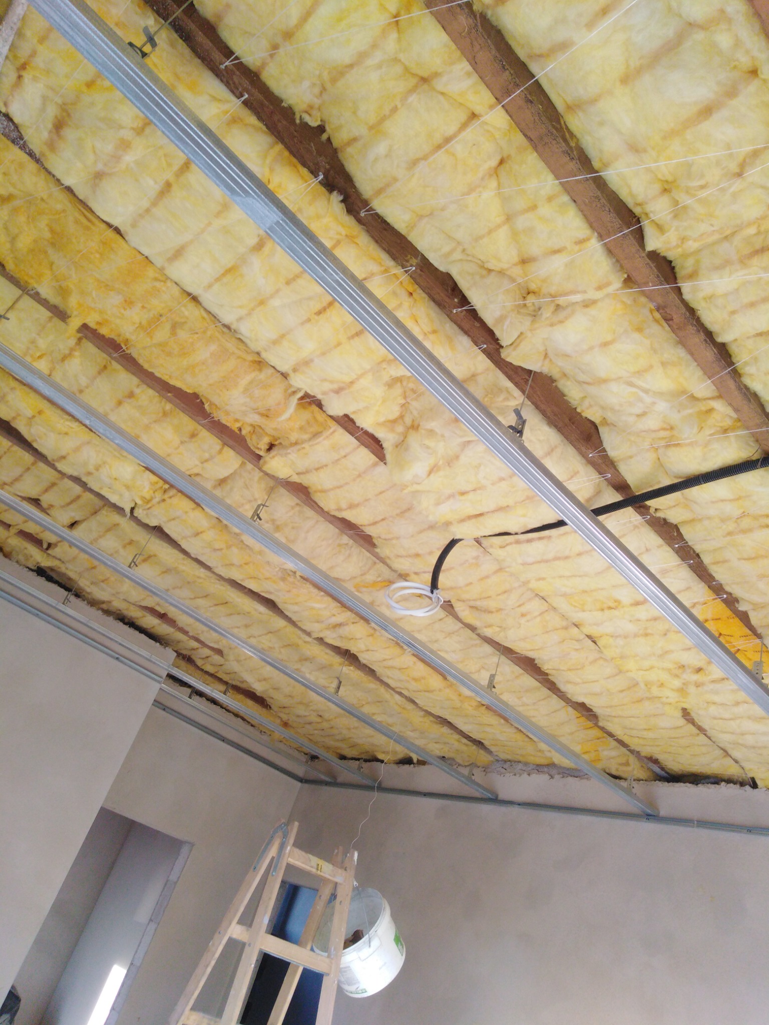 Ocieplenie poddasza z widoczną wełną mineralną między drewnianymi belkami i metalowymi profilami, przygotowane do montażu płyt gipsowo-kartonowych, drabina w rogu pomieszczenia.