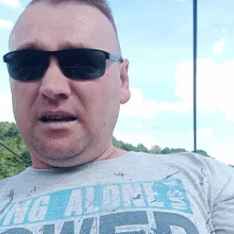 Autoportret mężczyzny w okularach przeciwsłonecznych na tle zieleni i nieba, z widoczną liną w tle.