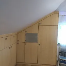 Jasna zabudowa meblowa w skosie poddasza, z szarymi, kwadratowymi uchwytami, widoczna drabina w rogu, parkiet na podłodze.