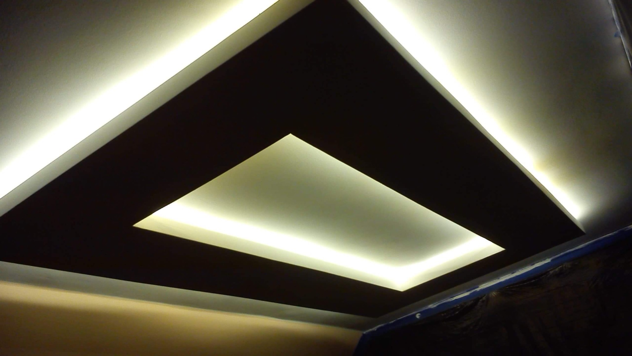 Nowoczesny sufit z podwieszanymi elementami pomalowanymi na ciemny brąz, oświetlony taśmami LED, widok z dołu.