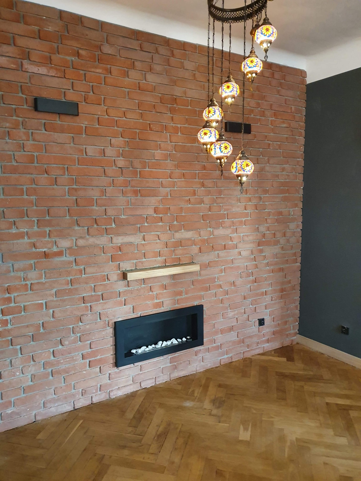 Ceglana ściana z wnęką na kominek, drewnianą półką i orientalną lampą wiszącą, parkiet w jodełkę.