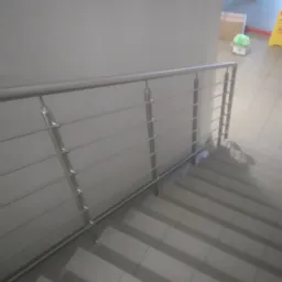 Wewnętrzne schody wyłożone szarymi płytkami z zamontowaną, prostą, metalową balustradą. W tle, na górze schodów, widoczna osoba w żółtej kamizelce.