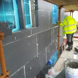 Pracownik w żółtej kurtce montuje czarne płyty izolacyjne na ścianie budynku, okna zabezpieczone folią, widoczne rusztowanie i narzędzia pracy.