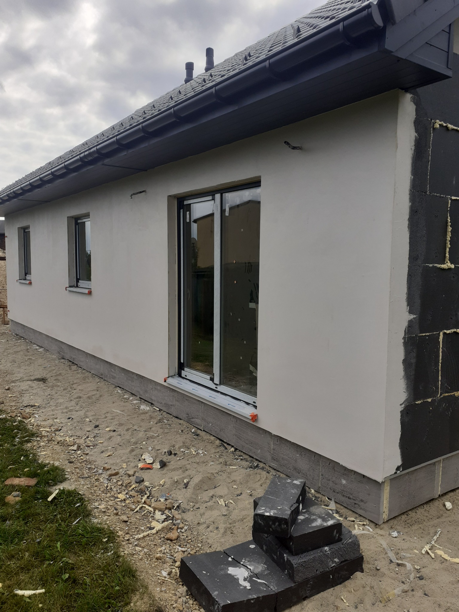 Fragment ocieplanego domu jednorodzinnego z widoczną elewacją po tynkowaniu, nieocieplonym fragmentem ściany z czarnym styropianem i zapasem czarnych bloczków na ziemi.