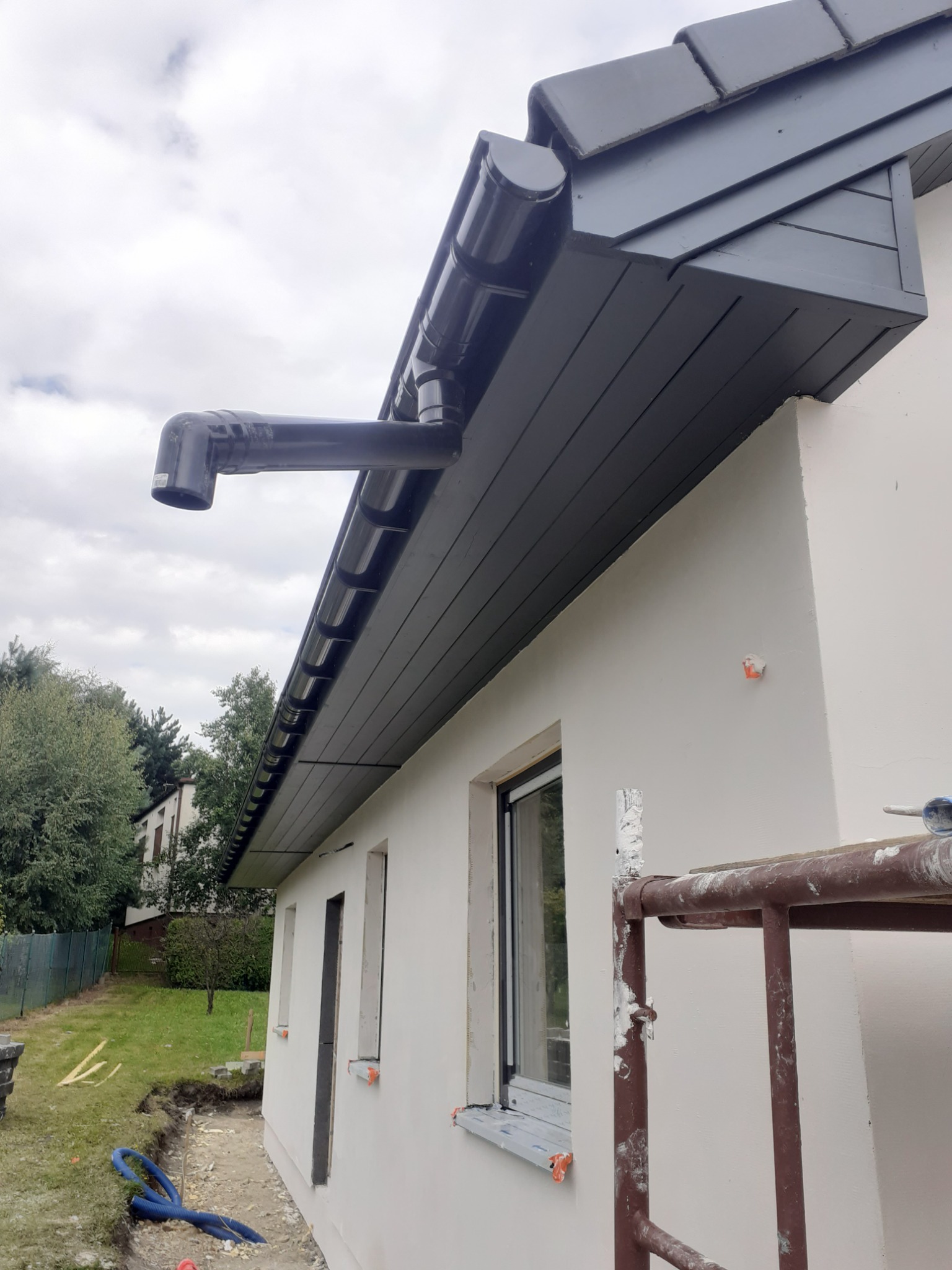 Nowa, ciemna rynna zamontowana na ocieplonym domu z widocznym rusztowaniem i przygotowanym terenem do zagospodarowania.