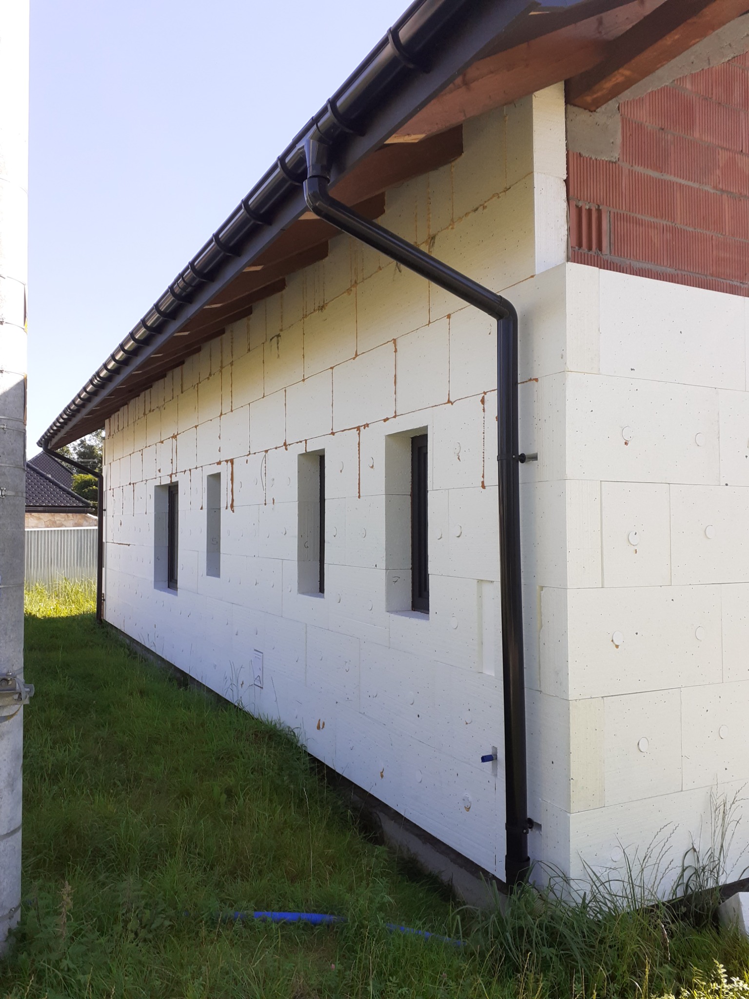 Boczny widok budynku w trakcie ocieplania, z widocznymi białymi płytami izolacyjnymi, czarną rynną i fragmentem nieocieplonej ściany z czerwonej cegły.