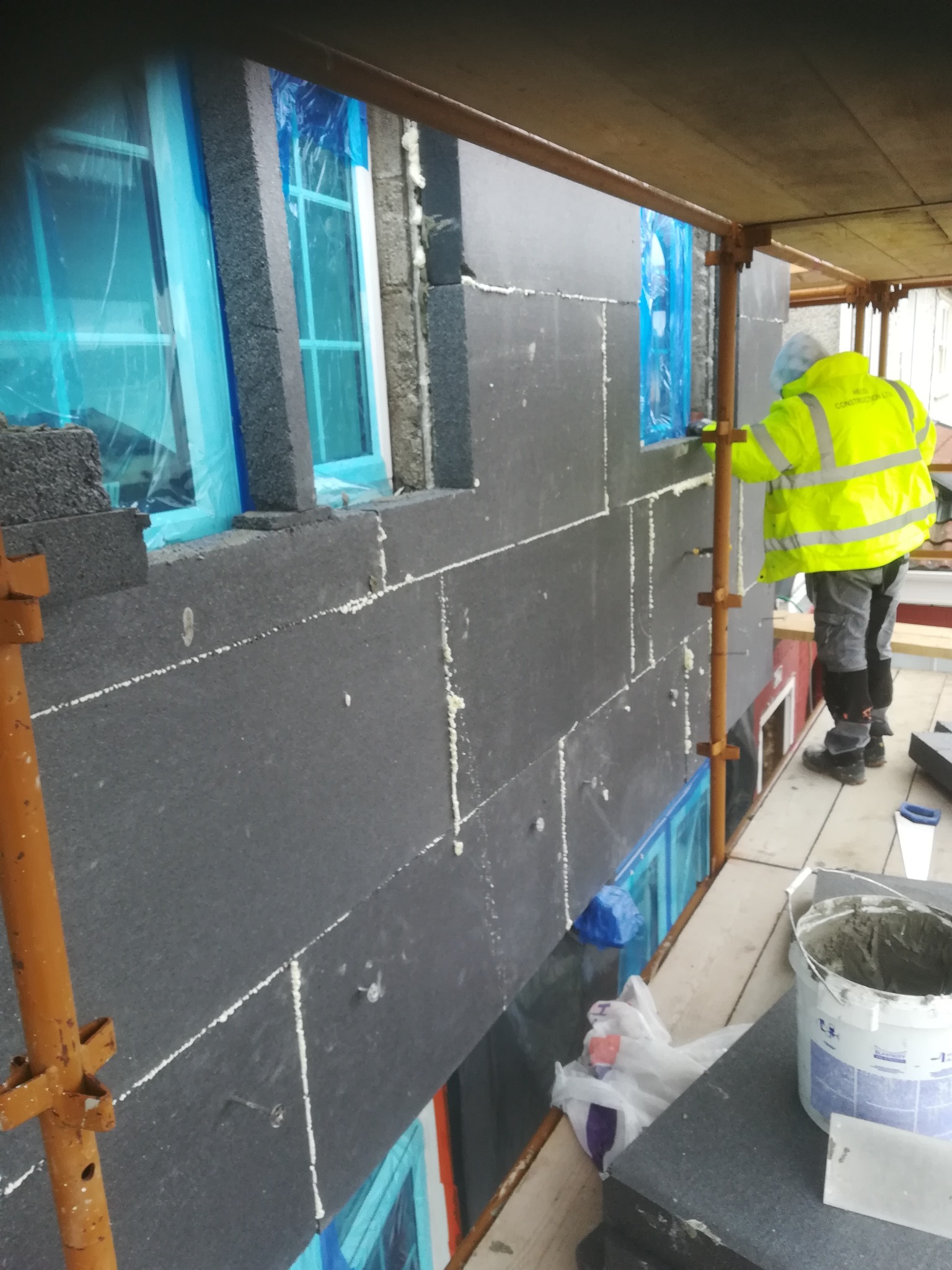 Pracownik w żółtej kurtce montuje czarne płyty izolacyjne na ścianie budynku, okna zabezpieczone folią, widoczne rusztowanie i narzędzia pracy.